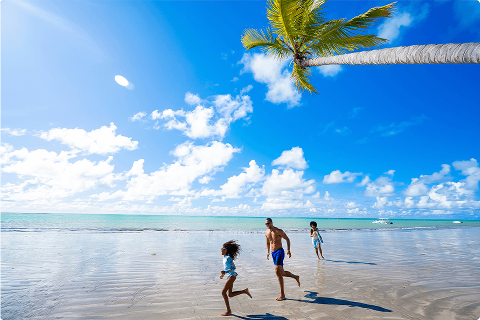 Pai correndo atrás da filha, com a mae no fundo sorrindo e brincando na praia, com um coqueiro inclinado, mar azul turques, ceu azul com poucas nuvens e alguns barcos ao fundo.