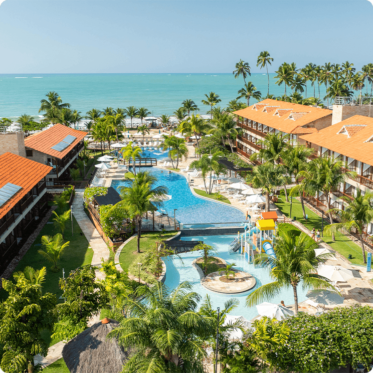 Vista aérea do resort Salinas Maceió, com apartamentos, piscinas, área de lazer infantil, coqueiros com mar e céu bem azul ao fundo.