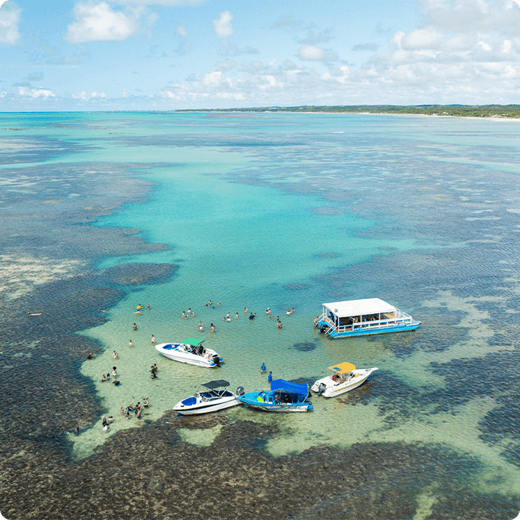 Vista aérea das piscinas naturais de Paripueira, em Maceió, com barcos e famílias nadando nas águas cristalinas com recifes de corais visíveis. Uma vegetação densa no fundo e céu com algumas nuvens. 
