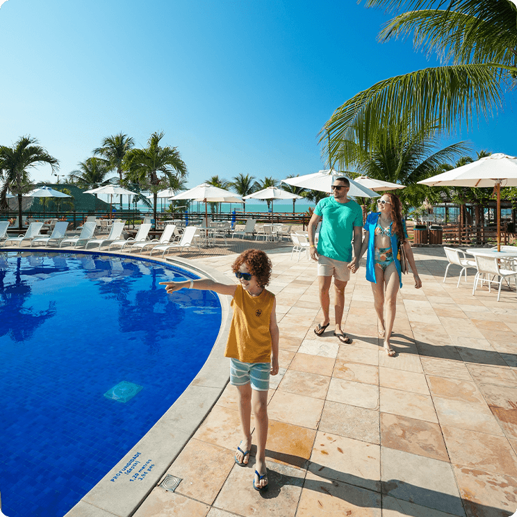 Família caminhando na beira da piscina do resort, com coqueiros, cadeiras de sol brancas ao fundo. Um bar do lado direito e um céu azul sem nuvem.