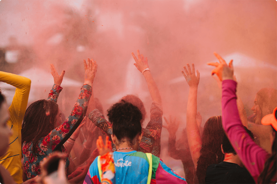 Várias crianças se divertindo em uma festa colorida com as mãos para cima e pó vermelho no ar. 