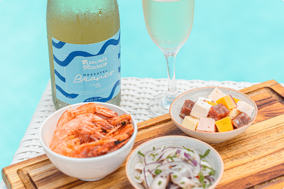 Moscatel Branco Salinas em garrafa com uma taça cheia ao lado e petiscos All Inclusive, como camarões e queijos, servidos à beira da piscina.
