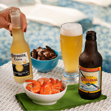 Comidas e bebidas do All Inclusive servidas ao lado da piscina, incluindo porção de camarões, fritas, quiches, um drink e cerveja gelada.