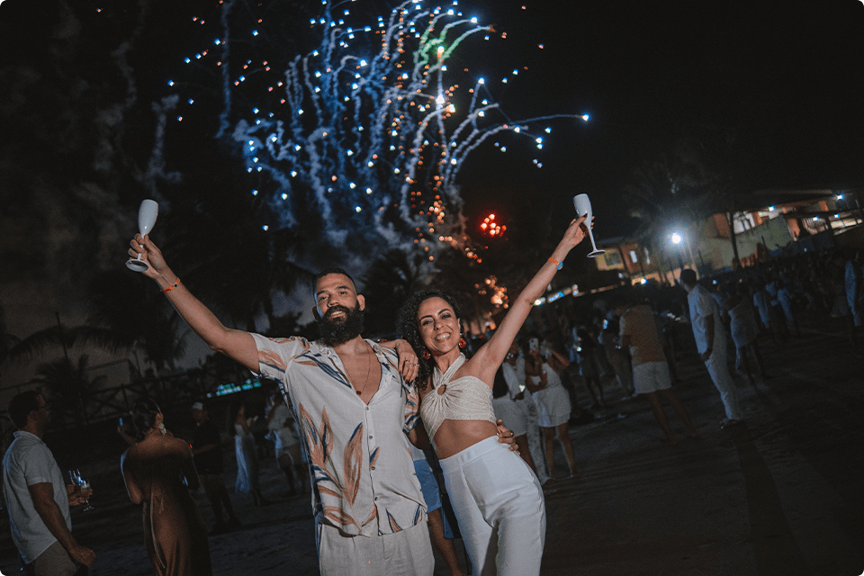 Casal com taças de champanhe comemorando o Réveillon na praia do resort Salinas Macéio, com fogos de artifício no céu e mais pessoas ao fundo curtindo também.