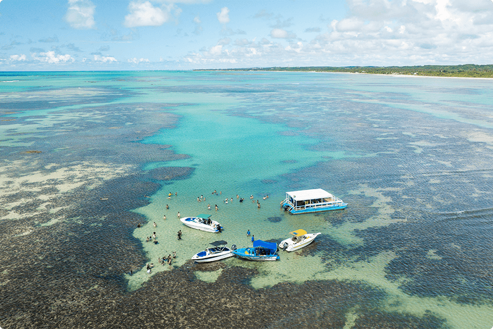 Vista aérea das piscinas naturais de Paripueira, em Maceió, com barcos e famílias nadando nas águas cristalinas com recifes de corais visíveis. Uma vegetação densa no fundo e céu com algumas nuvens. 