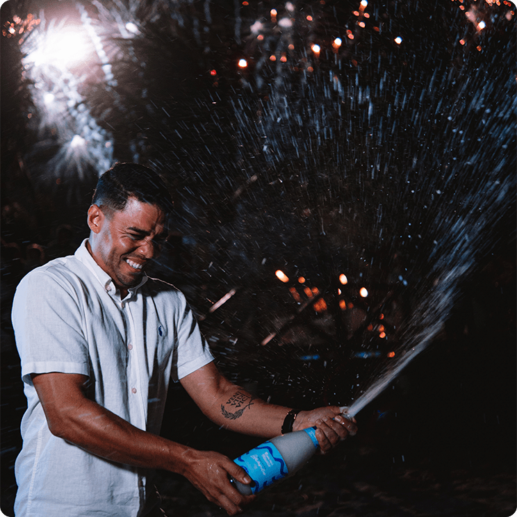 Homem estourando champanhe em celebração de Réveillon com fogos de artifício do céu ao fundo.