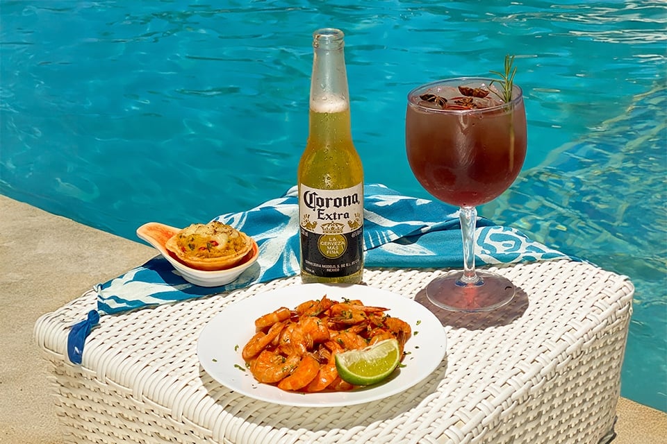 Cerveja long neck Corona Extra, taça de drink e petiscos sobre mesa à beira da piscina do Salinas Maceió.