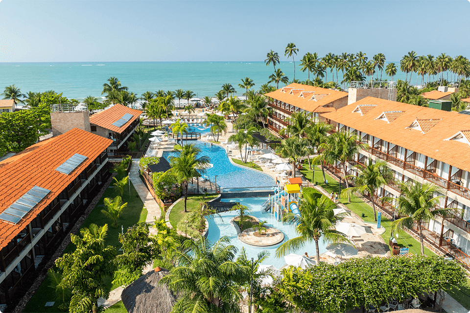Vista aérea do resort Salinas Maceió, com apartamentos, piscinas, área de lazer infantil, coqueiros com mar e céu bem azul ao fundo.