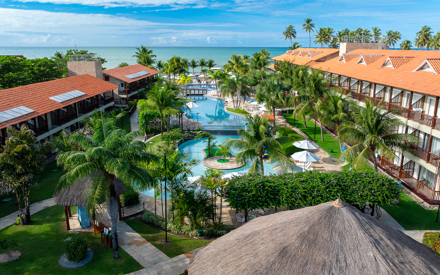 Vista aérea das piscinas e blocos de apartamentos do Salinas Maceió cercados por coqueirais sob céu azul. A família está na piscina infantil na área de lazer.