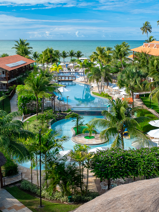 Vista aérea das piscinas e blocos de apartamentos do Salinas Maceió cercados por coqueirais sob céu azul. A família está na piscina infantil na área de lazer.