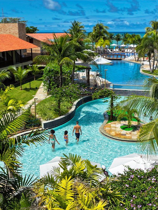 Estrutura completa pra família Vista aérea das piscinas e blocos de apartamentos do Salinas Maceió cercados por coqueirais sob céu azul. A família está na piscina infantil na área de lazer.