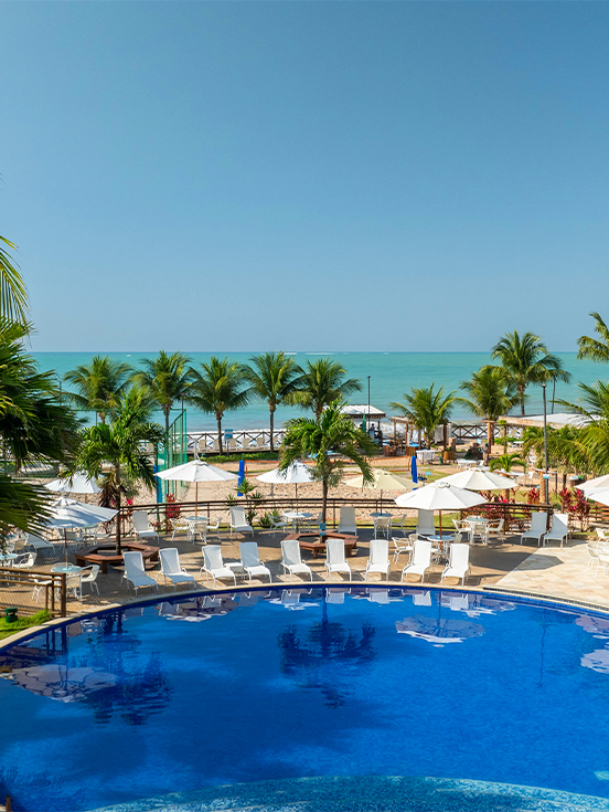 Área externa do Salinas Maceió com piscina ao centro, vegetação espalhada no resort, espreguiçadeira, guarda-sóis e ao fundo o mar.