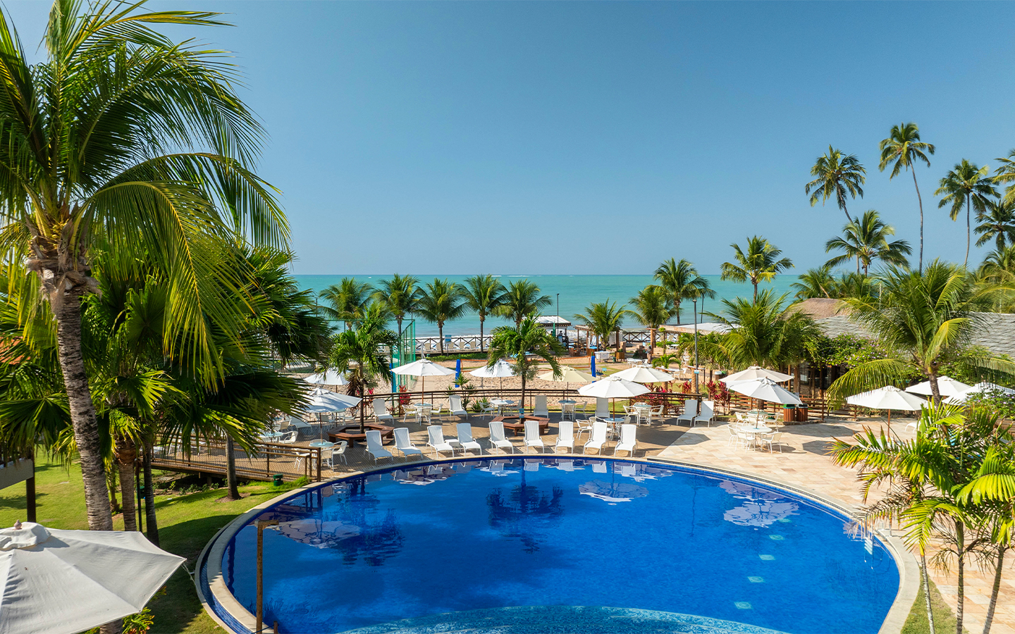 Área externa do Salinas Maceió com piscina ao centro, vegetação espalhada no resort, espreguiçadeira, guarda-sóis e ao fundo o mar.