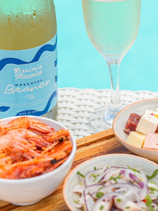 A garrafa de Moscatel do Salinas Maceió com taça ao lado. Petiscos como camarão, ceviche e queijos variados à beira da piscina.