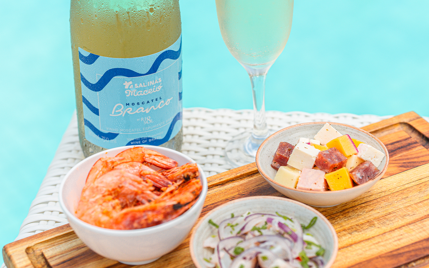A garrafa de Moscatel do Salinas Maceió com taça ao lado. Petiscos como camarão, ceviche e queijos variados à beira da piscina.