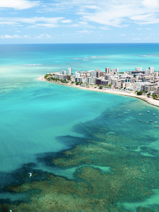 Praia de Ponta Verde em Maceió Vista aérea da Praia de Ponta Verde em Maceió com mar verde-esmeralda, recifes de corais, barcos ancorados e a orla urbana ao fundo sob céu azul.