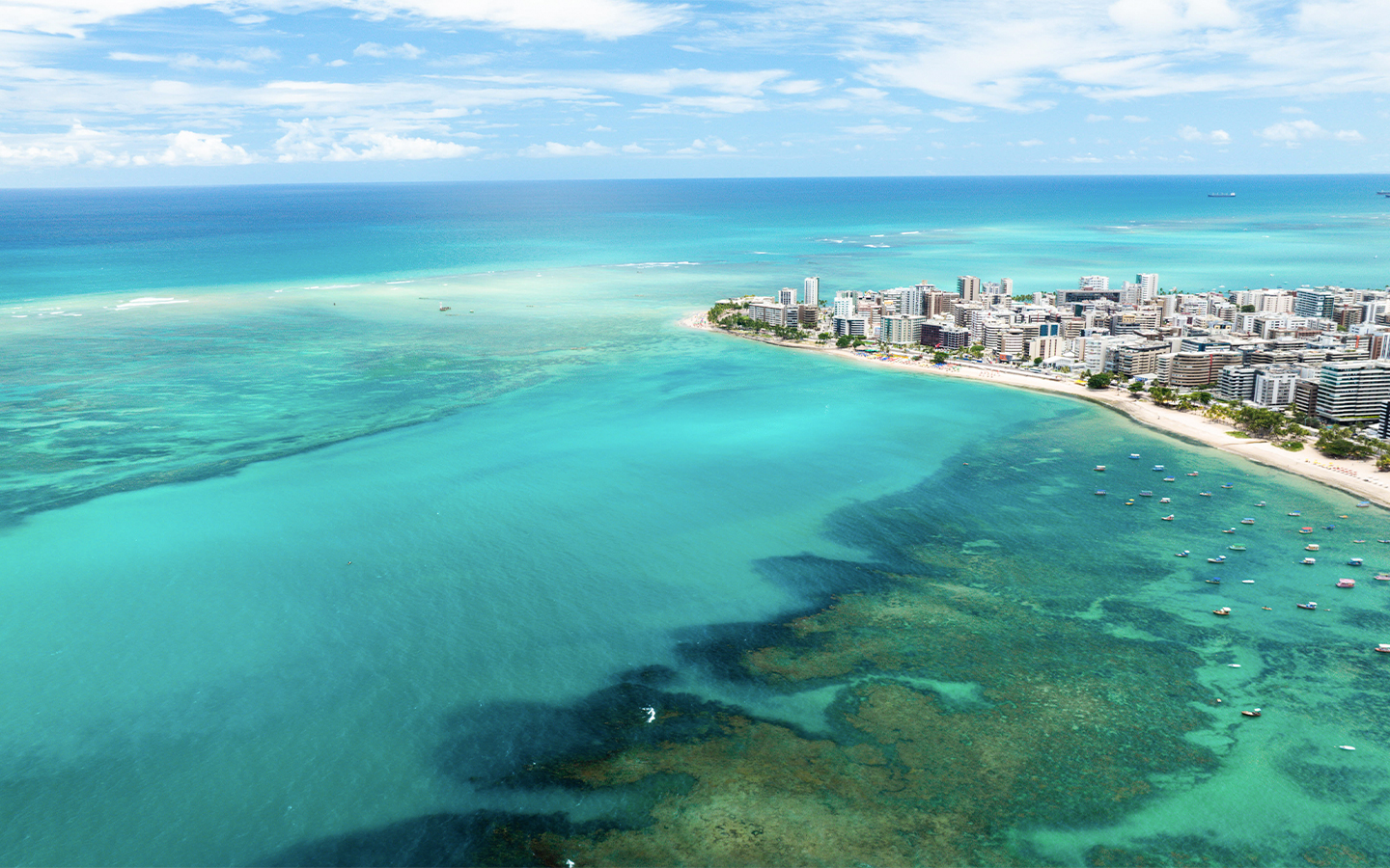 Praia de Ponta Verde em Maceió Vista aérea da Praia de Ponta Verde em Maceió com mar verde-esmeralda, recifes de corais, barcos ancorados e a orla urbana ao fundo sob céu azul.