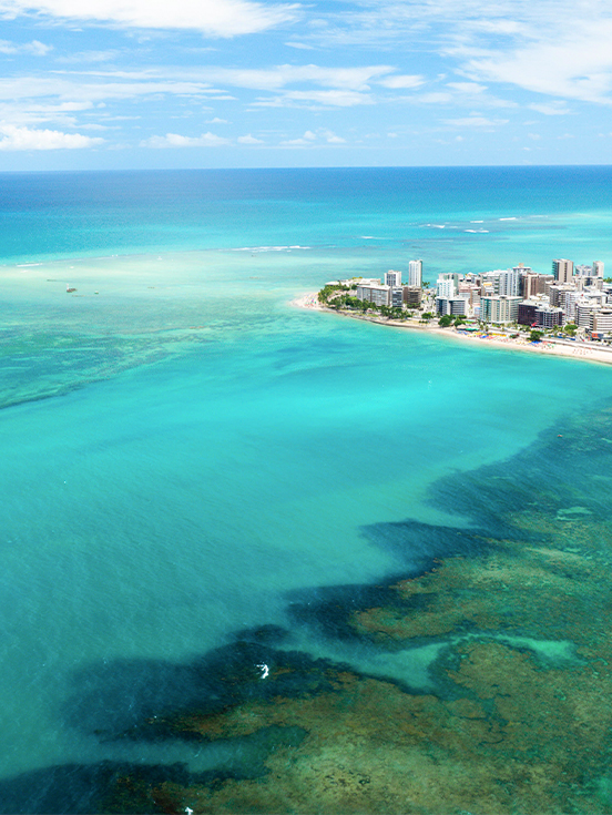 Vista aérea da Praia de Ponta Verde em Maceió com mar verde-esmeralda, recifes de corais, barcos ancorados e a orla urbana ao fundo sob céu azul.