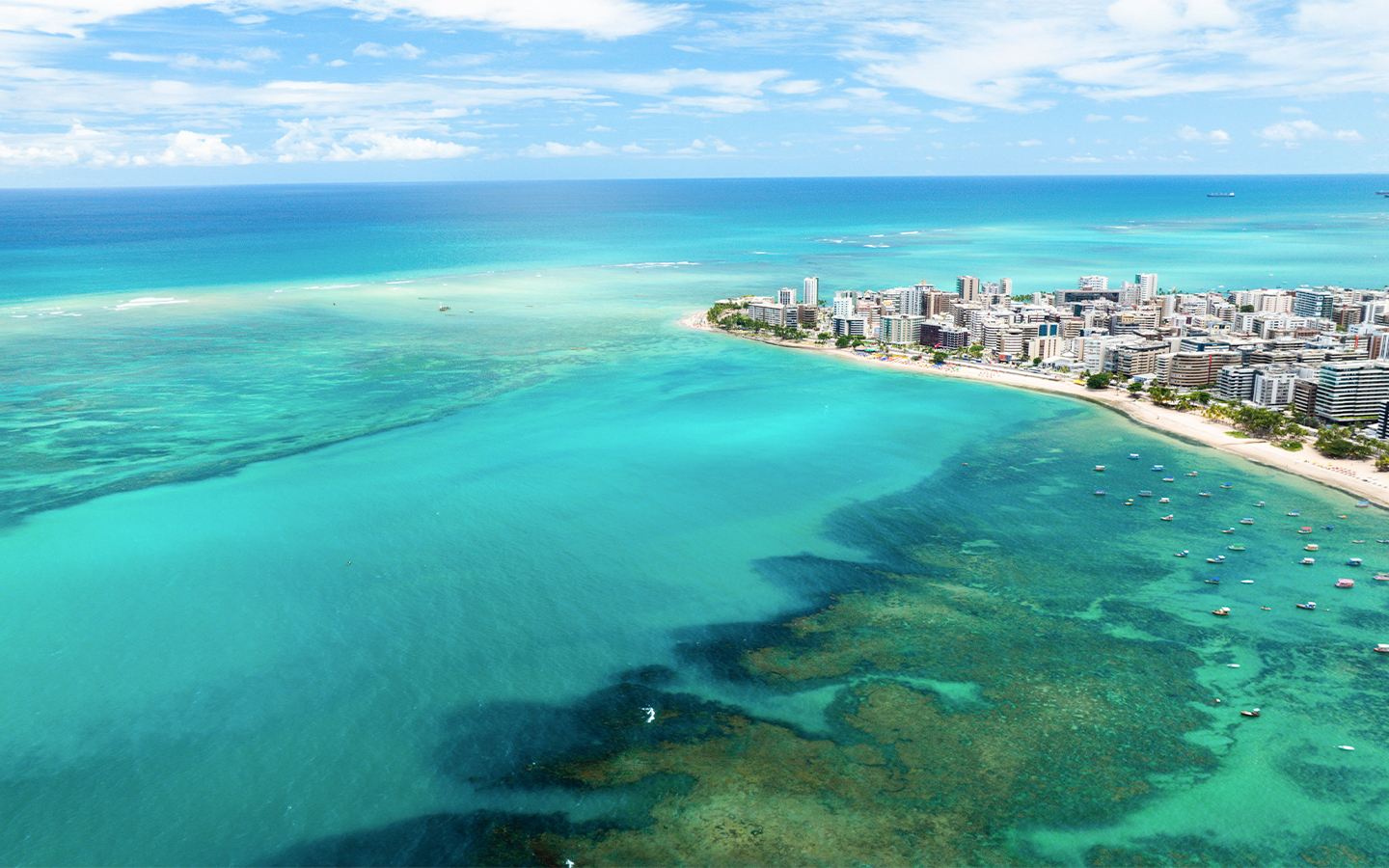 Vista aérea da Praia de Ponta Verde em Maceió com mar verde-esmeralda, recifes de corais, barcos ancorados e a orla urbana ao fundo sob céu azul.
