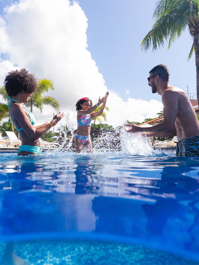 Família se divertindo na piscina do Salinas Maceió com pai, mãe e filha jogando água pra cima. Ao redor toda a estrutura do resort, com espreguiçadeiras, coqueiros e guarda-sóis.