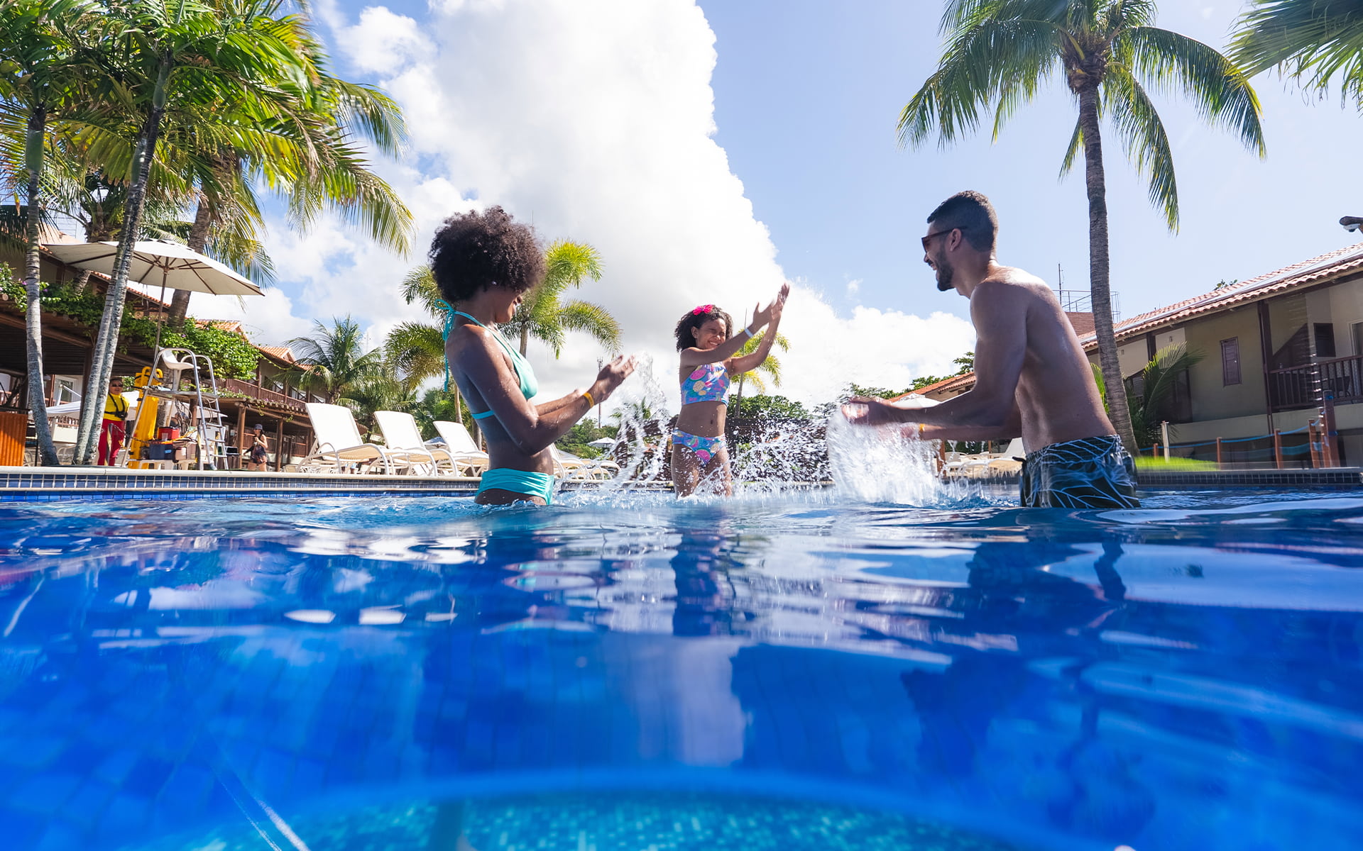 Família se divertindo na piscina do Salinas Maceió com pai, mãe e filha jogando água pra cima. Ao redor toda a estrutura do resort, com espreguiçadeiras, coqueiros e guarda-sóis.