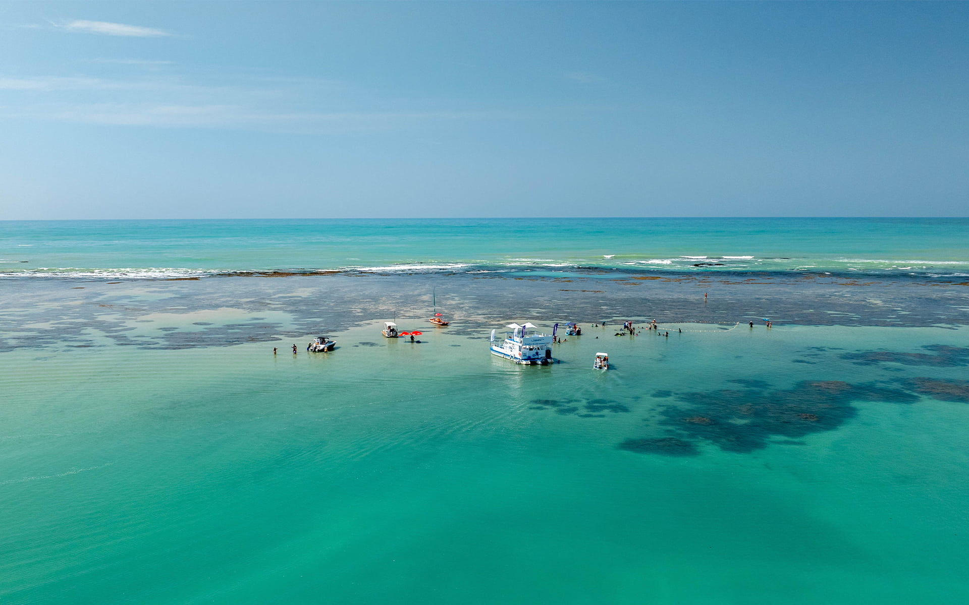 Vista aérea da Praia de Pajuçara com mar cristalino, recifes de corais visíveis e algumas embarcações atracadas nas piscinas naturais.