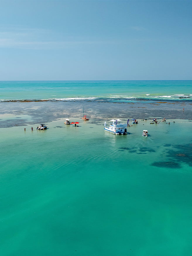 Vista aérea da Praia de Pajuçara com mar cristalino, recifes de corais visíveis e algumas embarcações atracadas nas piscinas naturais.