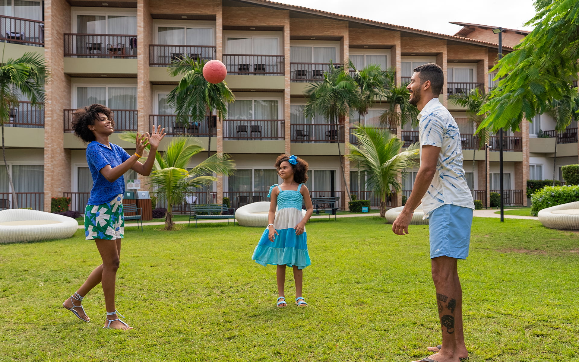 Família brincando de bola no gramado ao ar livre no Salinas Maceió, com toda estrutura dos apartamentos ao fundo.