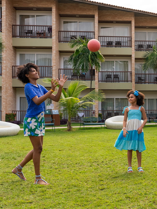 Família brincando de bola no gramado ao ar livre no Salinas Maceió, com toda estrutura dos apartamentos ao fundo.