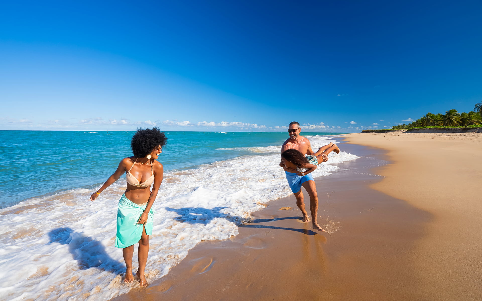 Família caminhando na Praia do Gunga com mãe a frente, e pai brincando de avião com a filha na faixa de areia com dia ensolarado.