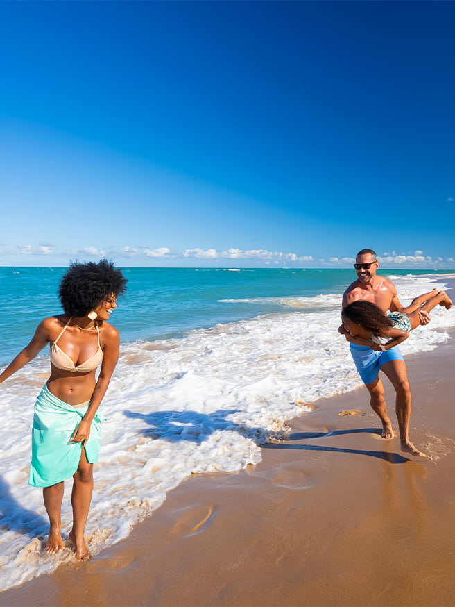 Família caminhando na Praia do Gunga com mãe a frente, e pai brincando de avião com a filha na faixa de areia com dia ensolarado.