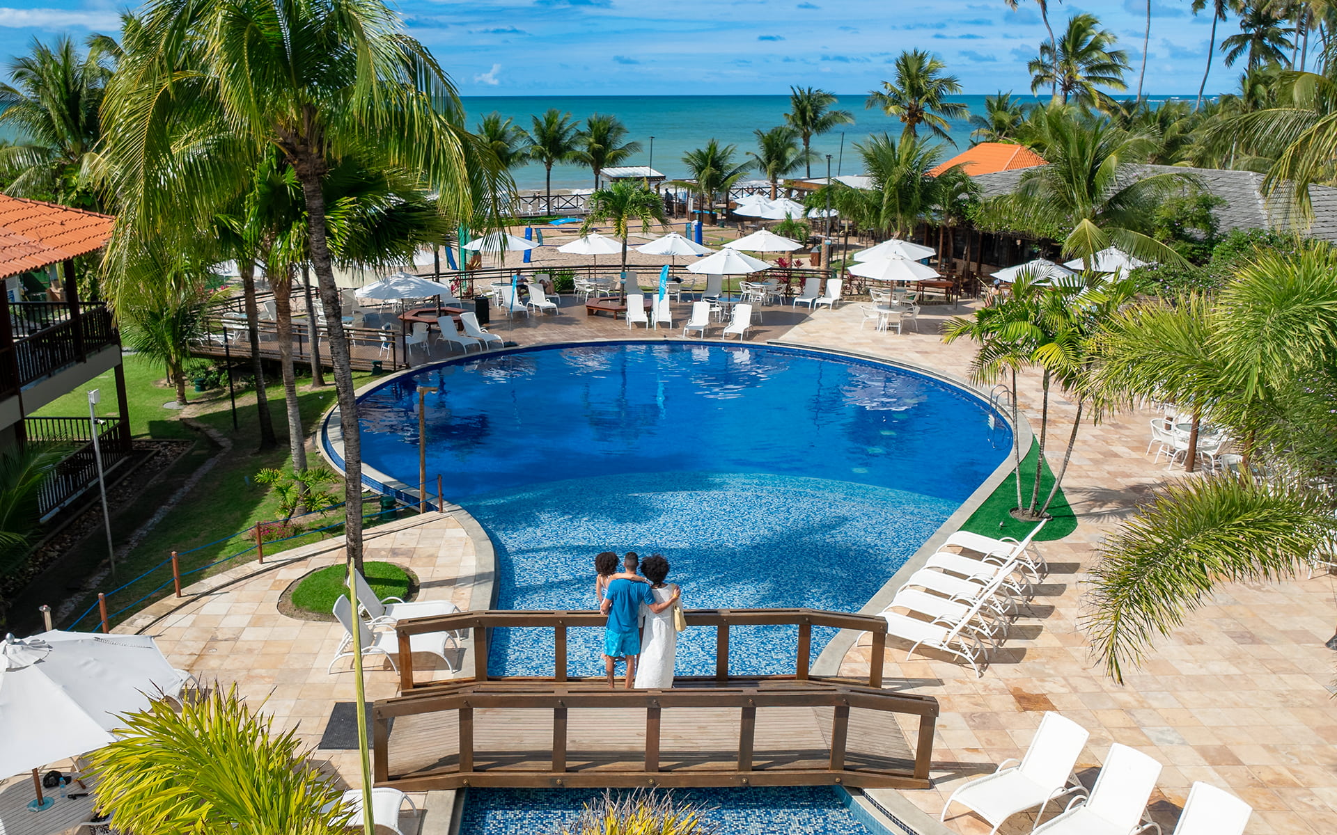 Piscina redonda, com ponte de madeira passando por cima dela. Na ponta há uma família de pai, mãe e filha observando o horizonte do resort e praia do Salinas Maceió cercada por coqueiros e áreas de lazer.