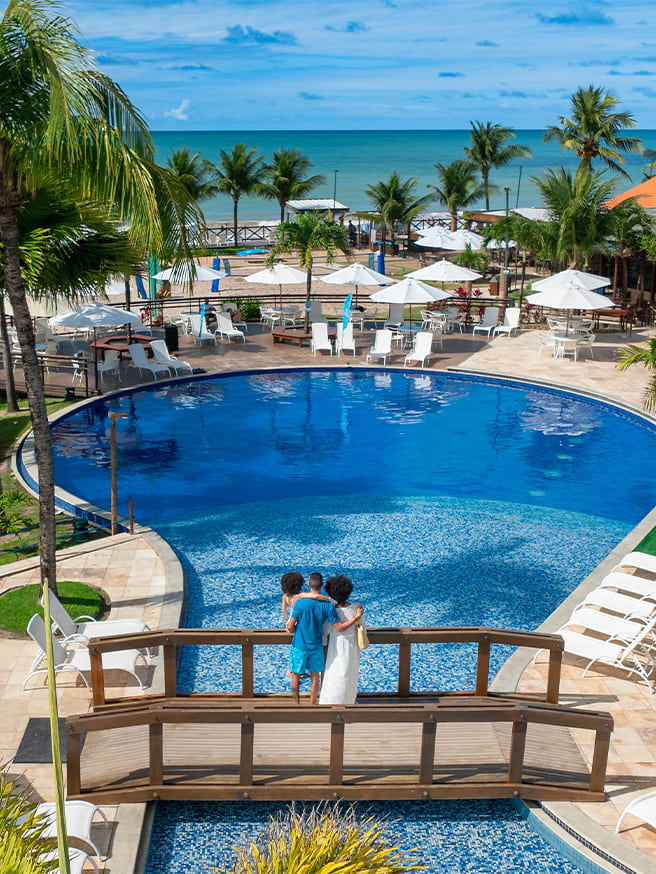 Piscina redonda, com ponte de madeira passando por cima dela. Na ponta há uma família de pai, mãe e filha observando o horizonte do resort e praia do Salinas Maceió cercada por coqueiros e áreas de lazer.