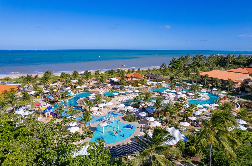 Vista aérea do complexo de piscinas do Salinas Maceió, é possível ver brinquedos infantis, guarda-sois, e pessoas se divertindo. Com muitas árvores ao redor, e ao fundo a praia de Maragogi.
