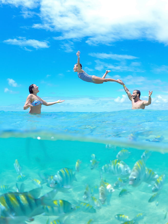 Família se divertindo nas piscinas naturais de Paripueira. Com foto meia submersa, na parte inferior está abaixo d'água com peixinhos e acima, o pai jogando a filha pra cima em direção a mãe com dia ensolarado.
