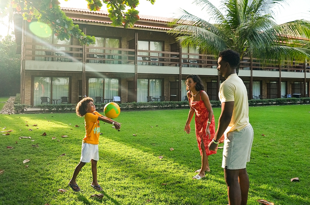 Família jogando volei na área de grama do Salinas Maragogi, em frente aos apartamentos. O dia está ensolarado no fim de tarde. A criança está jogando a bola pra cima.