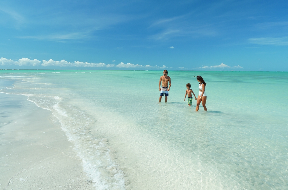 A Praia de Antunes com águas cristalinas e azul turquesa, com família no rosa, a areia é branca e o céu está quase sem nuvens.
