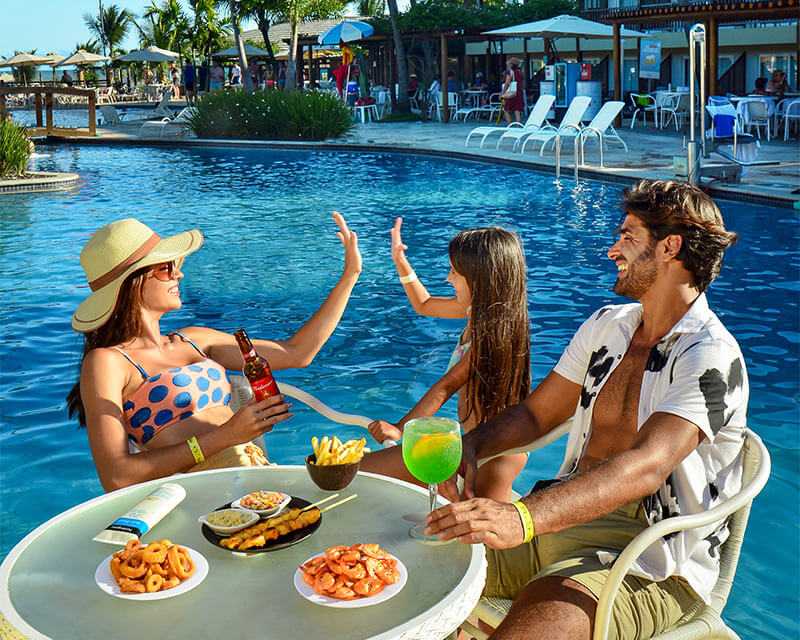 Homem, mulher e filha felizes curtindo o Salinas Maceió em mesa perto da piscina.