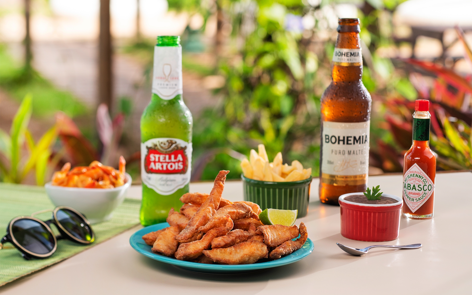 resort-all-inclusive Cerveja long neck Stella Artois e Bohemia Puro Malte, ao lado de uma pimenta Tabaco, e petiscos, camarão alho e óleo, camarão empanado, batata frita, disponíveis no resort All Inclusive Salinas Maceió sobre tábuas de madeira em mesa branca à beira da piscina.