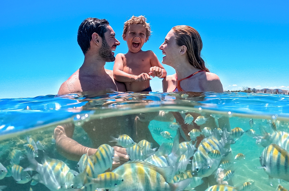 O contato com a natureza é único Foto subaquática com família dentro do mar, com água até os ombros, o pai está com seu filho pequeno no colo e a mãe olhando pra eles sorrindo. Há inúmeros peixinhos listrados ao redor da família, o céu está azul sem nuvens.