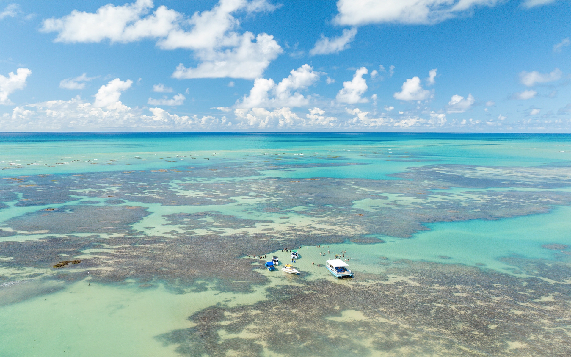 piscinas-naturais-paripueira Vista aérea das piscinas naturais de Paripueira, água azul turquesa com jangadas e barcos em passeio. Possível ver a imensidão dos recifes de corais.