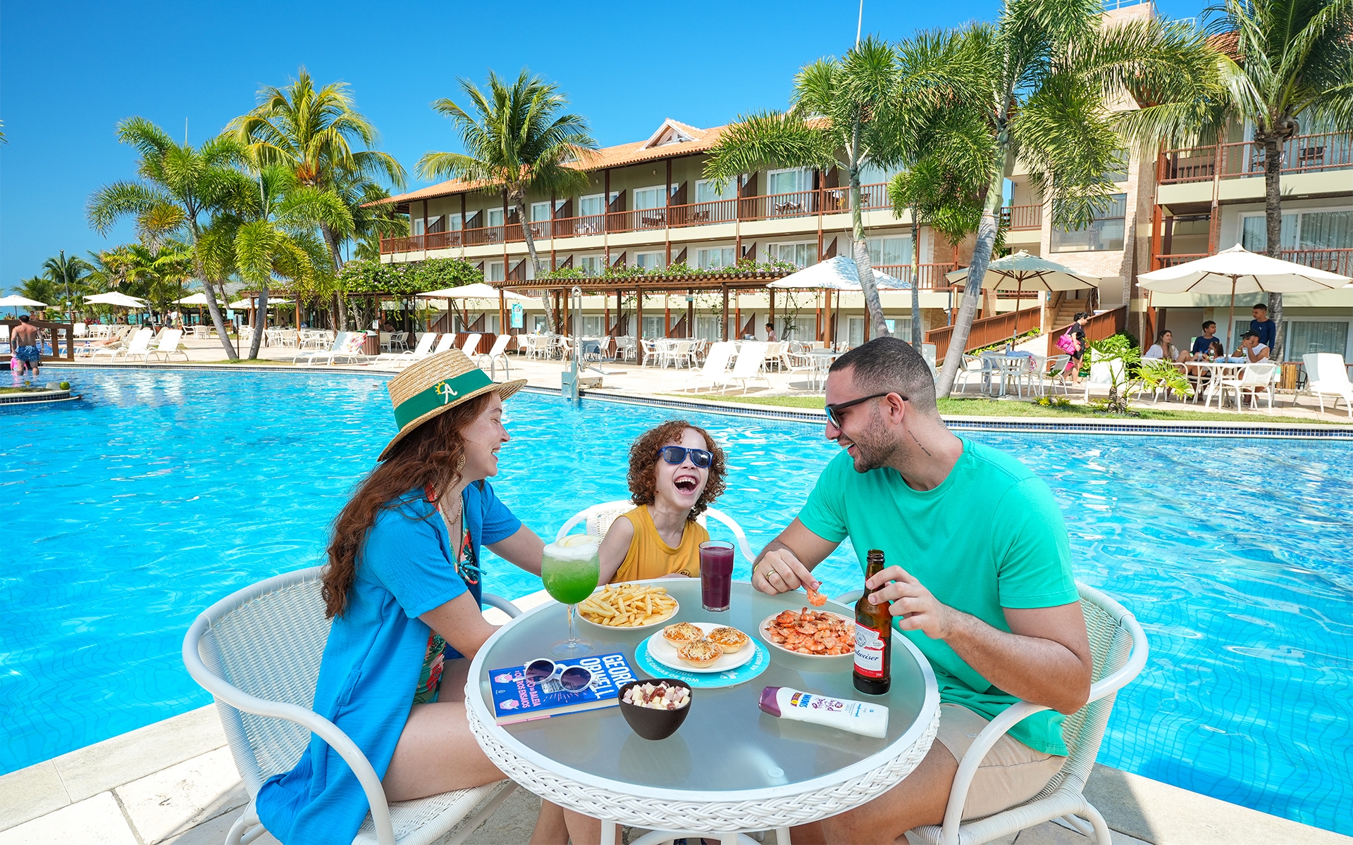 resort-familia-de-praia Família desfruta de petiscos e drinks em mesa à beira da piscina no Salinas Maceió, ao fundo as piscinas e bloco de apartamentos com varanda pra área de lazer. O dia está ensolarado e sem nuvens.