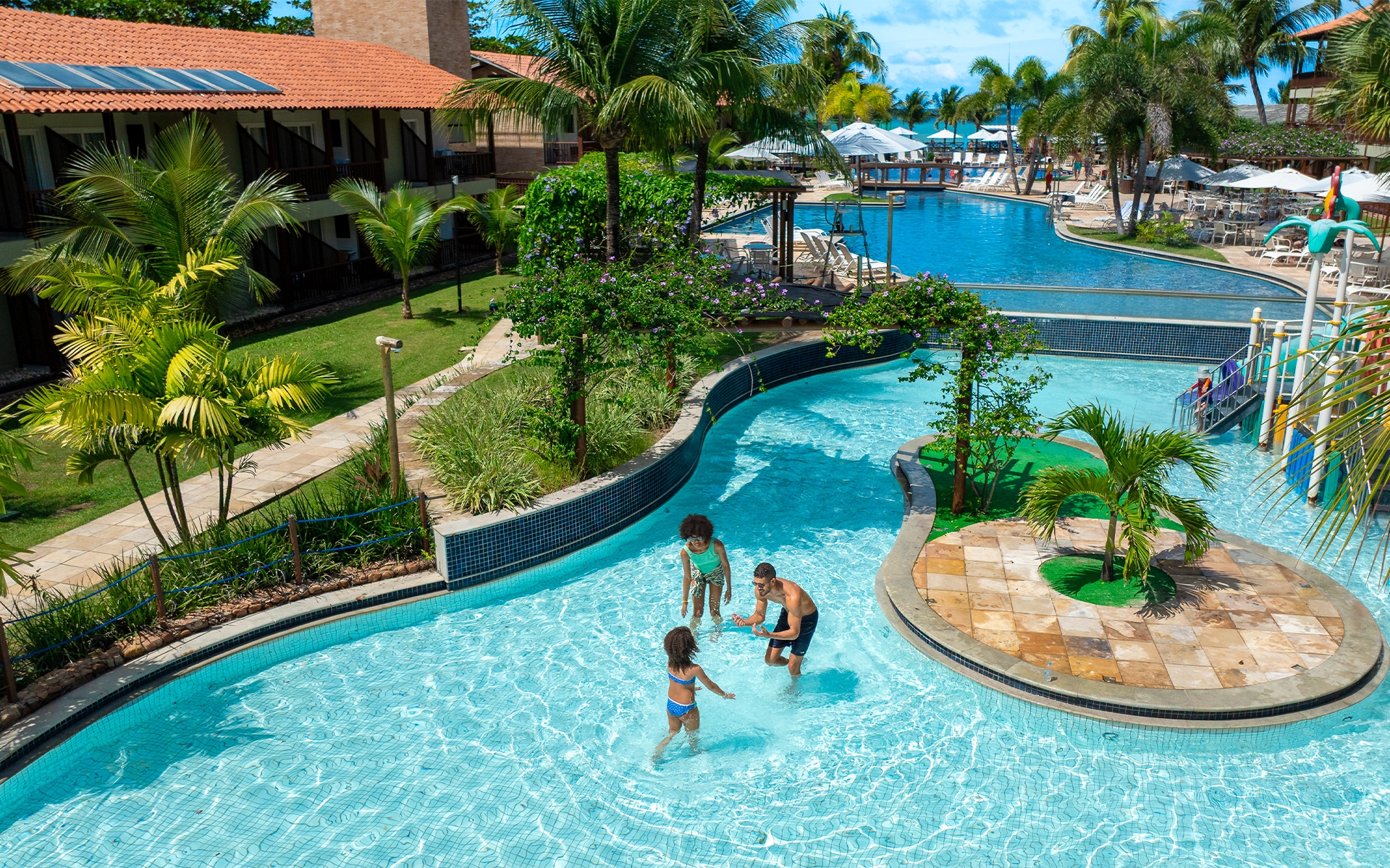 estrutura-completa-salinas-maceió Família na piscina infantil do Salinas Maceió, em frente às acomodações do resort All Inclusive à beira-mar da Praia de Ipioca. Ao redor da piscina há guarda-sóis, espreguiçadeiras e inúmeros coqueiros.