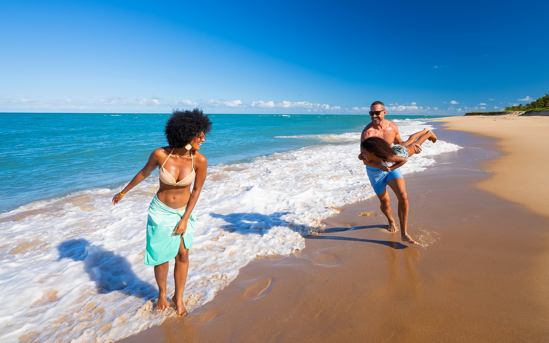 praia-do-gunga Família na praia do Gunga, com faixa de areia clara e mar cor azul. Família brinca perto da água, pai carregando filha como se fosse avião e mãe sorri olhando pra eles.