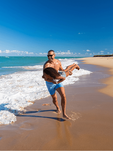 praia-do-gunga-mobile Pai e filha brincando na praia, homem segurando a criança com braços abertos na orla, mar azul turquesa e céu limpo.