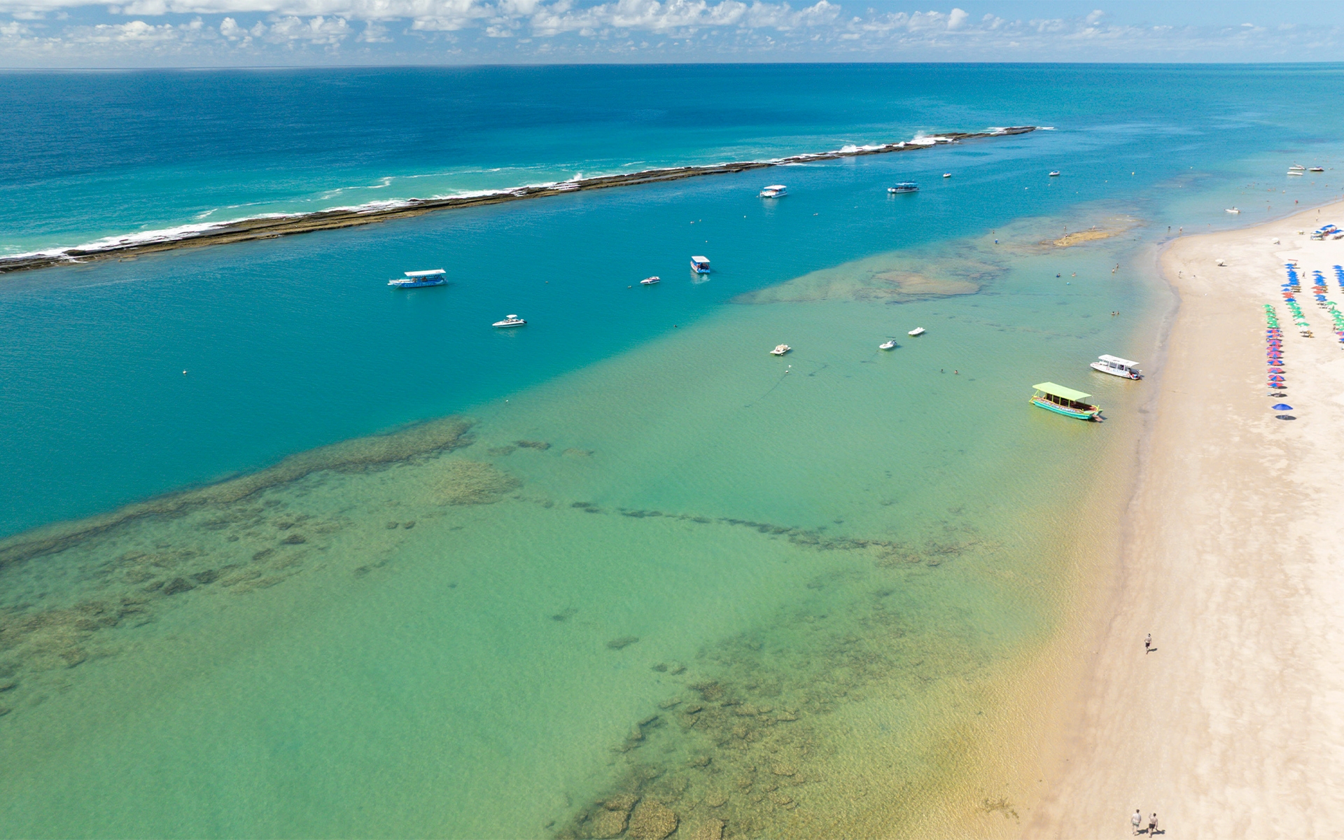 praia-do-frances Vista aérea da Praia do Francês, com maré baixa. Possível ver a barreira de corais, com alguns barcos na parte rasa. Na faixa de areia, algumas barracas e guarda-sóis coloridos.