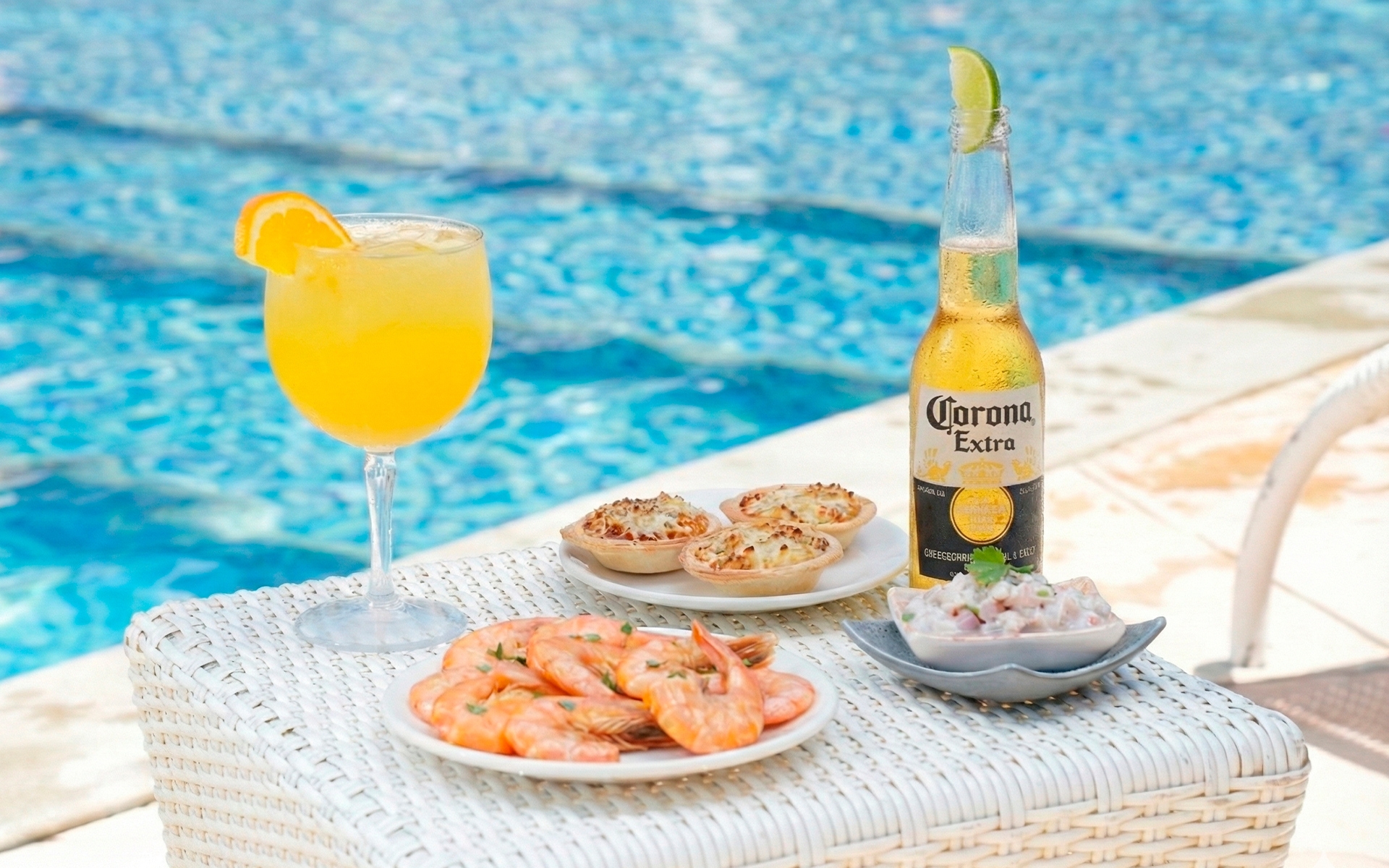 all-inclusive-salinas-maceió Drink amarelo com rodela de laranja na borda da taça, ao lado de prato de camarões, empadas e ceviche e uma long neck de Corona Extra com pedaço de limão na boca da garrafa. E ao fundo a piscina.