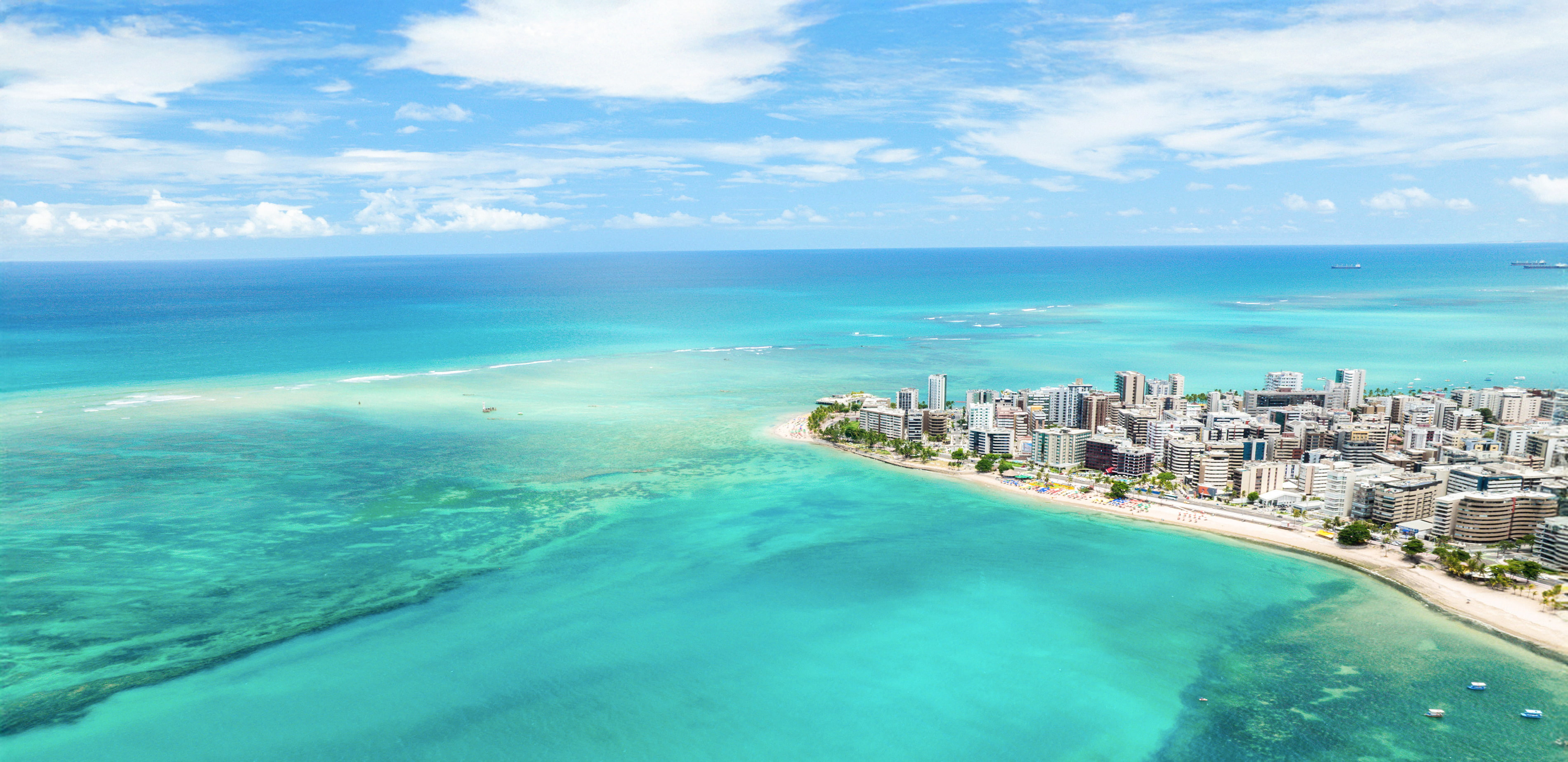 Vista aérea da praia de Ponta Verde em Maceió. O mar tem cor azul turquesa e é possível ver corais ao fundo por água ser cristalina. Ao lado direito a orla da cidade com prédios.