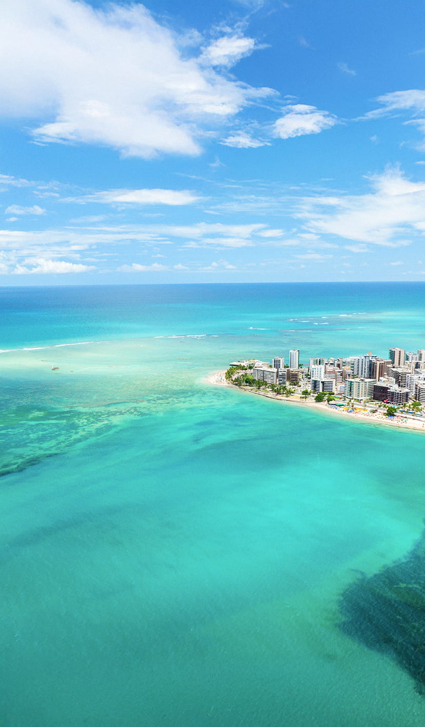 Vista aérea da praia de Ponta Verde em Maceió. O mar tem cor azul turquesa e é possível ver corais ao fundo por água ser cristalina. Ao lado direito a orla da cidade com prédios.