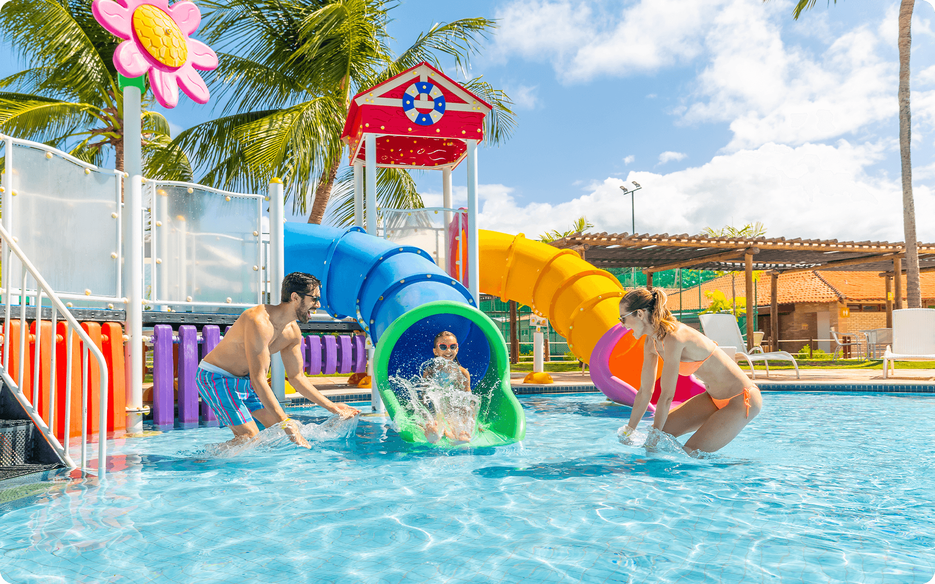 Família brincando nas piscinas, com filha descendo o toboágua infantil enquanto o pai e mãe jogam água pra cima, o dia é ensolarado. 